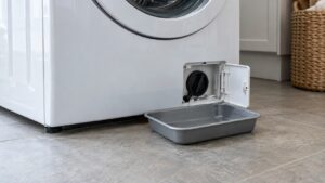 The lower front corner of a front-loading washer with a small service flap open and a shallow pan on the floor
